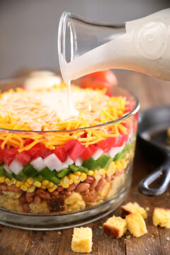 Layered Cornbread Salad in clear glass bowl - buttermilk ranch being poured over the top