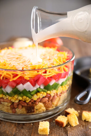 Layered Cornbread Salad in clear glass bowl - buttermilk ranch being poured over the top